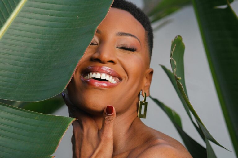 A vibrant and happy woman with short hair laughing behind lush green leaves, showcasing natural beauty.